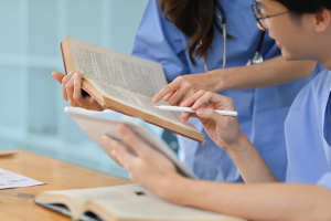 Two medical students studying together using a textbook and a tablet in a classroom setting, wearing blue scrubs.